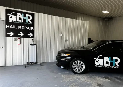 Black car parked indoors next to a large “Hail Repair” sign with directional arrows and a BYR logo.