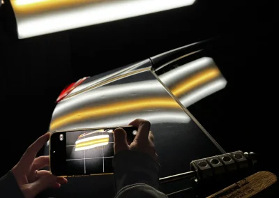 Hands taking a photo of a car's shiny surface reflecting yellow and white lights in a dark setting.