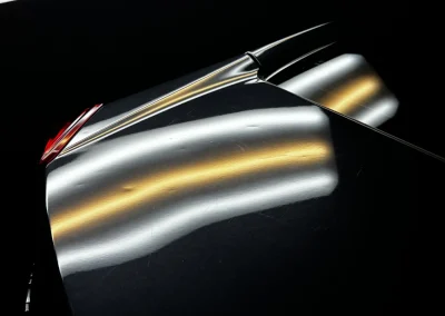Close-up of a dark car hood with visible dents and reflections of overhead lights.