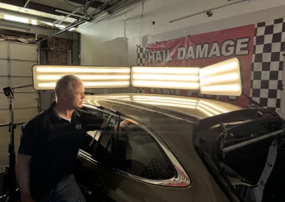 A man inspects a car under bright lights next to a red "Hail Damage" sign in a garage.