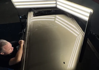 A person repairs dents on a car roof under bright inspection lights in an indoor workshop.