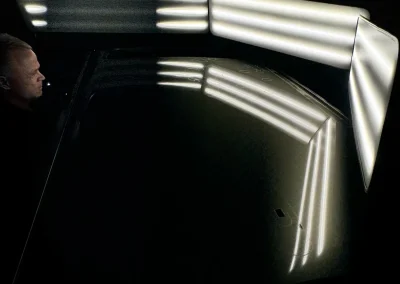 A person inspects a shiny, dark surface reflecting bright, horizontal LED lights in a dark room.