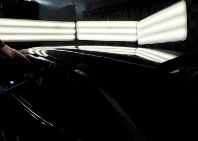 A row of bright lights reflects on the shiny black roof of a car in a dimly lit garage.