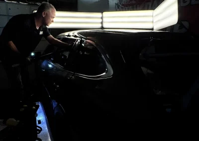 A man inspects a car's roof under bright panel lights in a dark auto repair workshop.