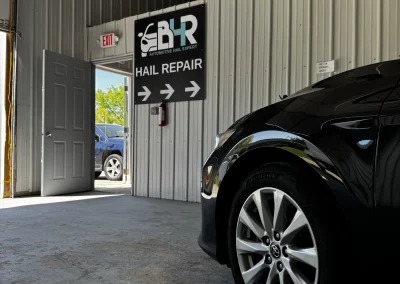 A black car parked inside a hail repair shop, with an open door showing a car outside.