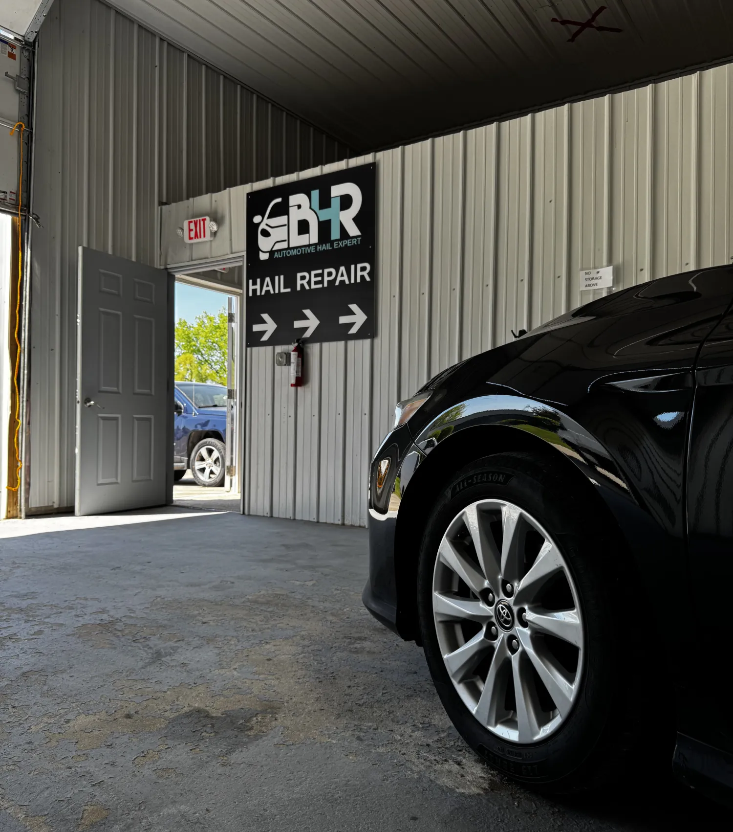 A black car parked inside a hail repair shop, with an open door showing a car outside.