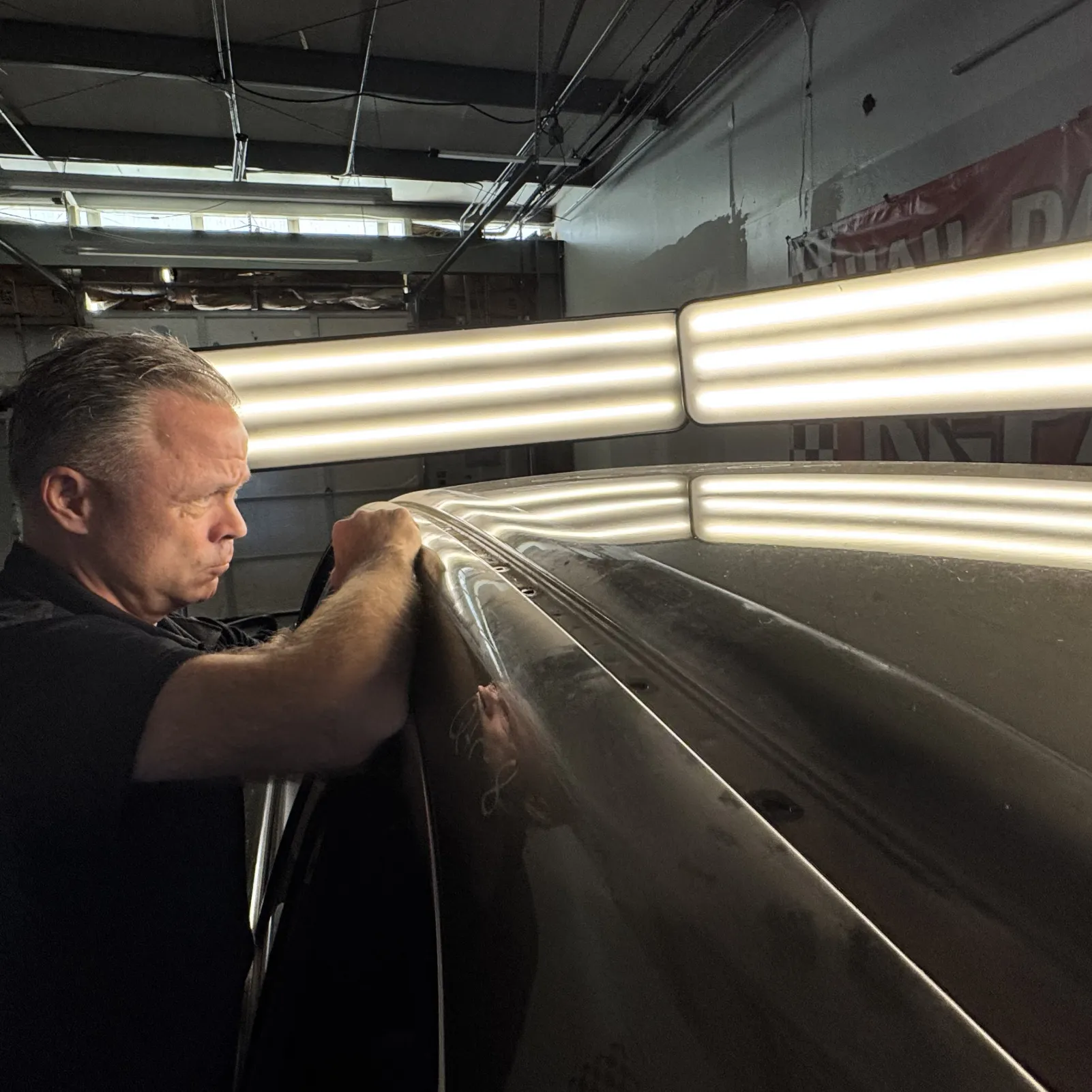 A man repairs a car roof under bright lights in a garage, with reflections visible on the car's surface.