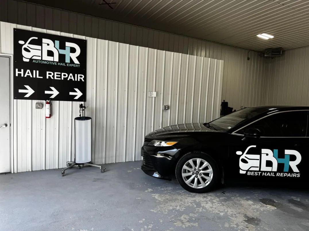 Black car with "Best Hail Repairs" logo parked in a garage next to a "Hail Repair" sign with arrows.