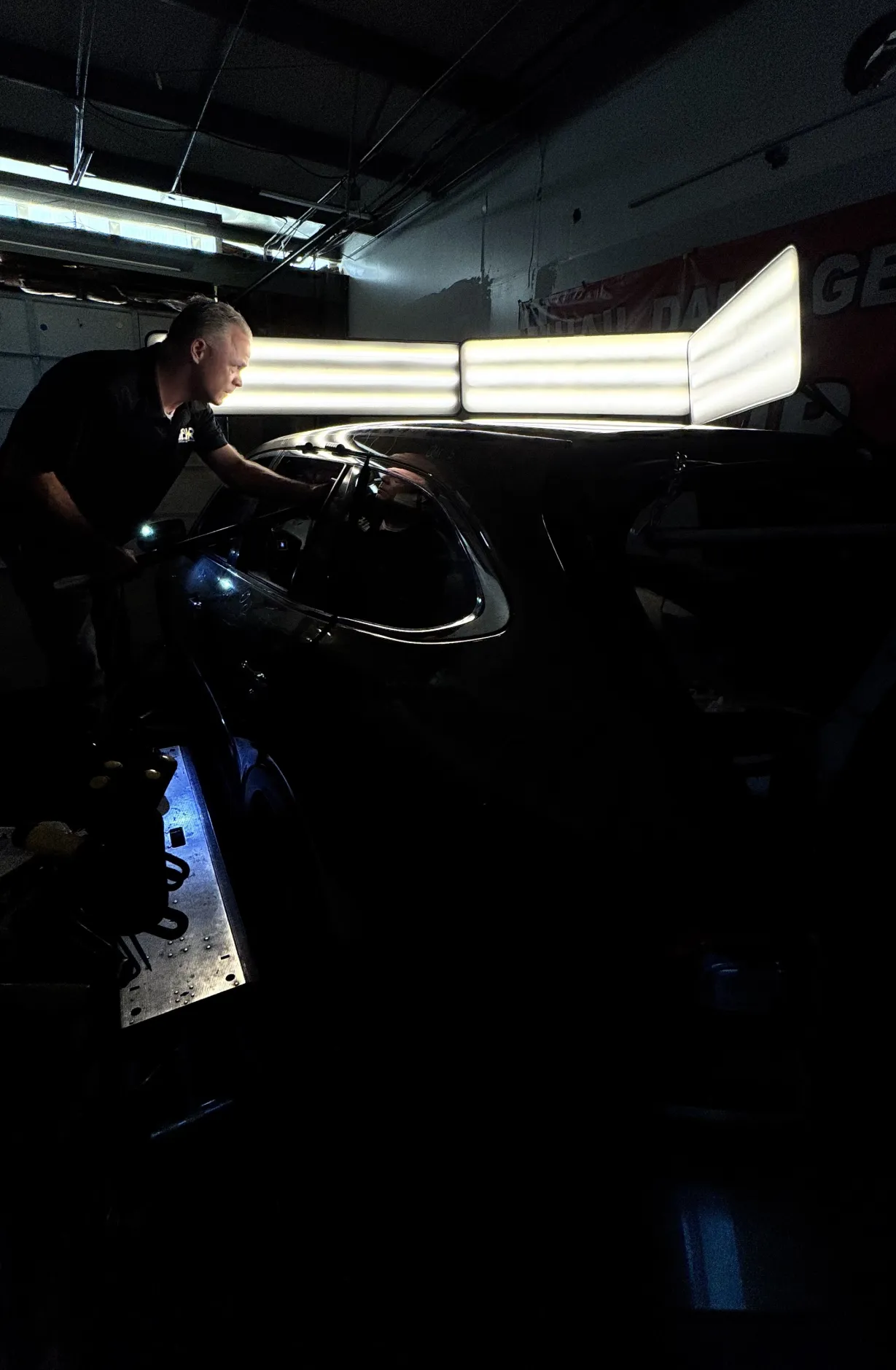 A man inspects a car under bright lights in a dimly lit garage, using a flashlight.
