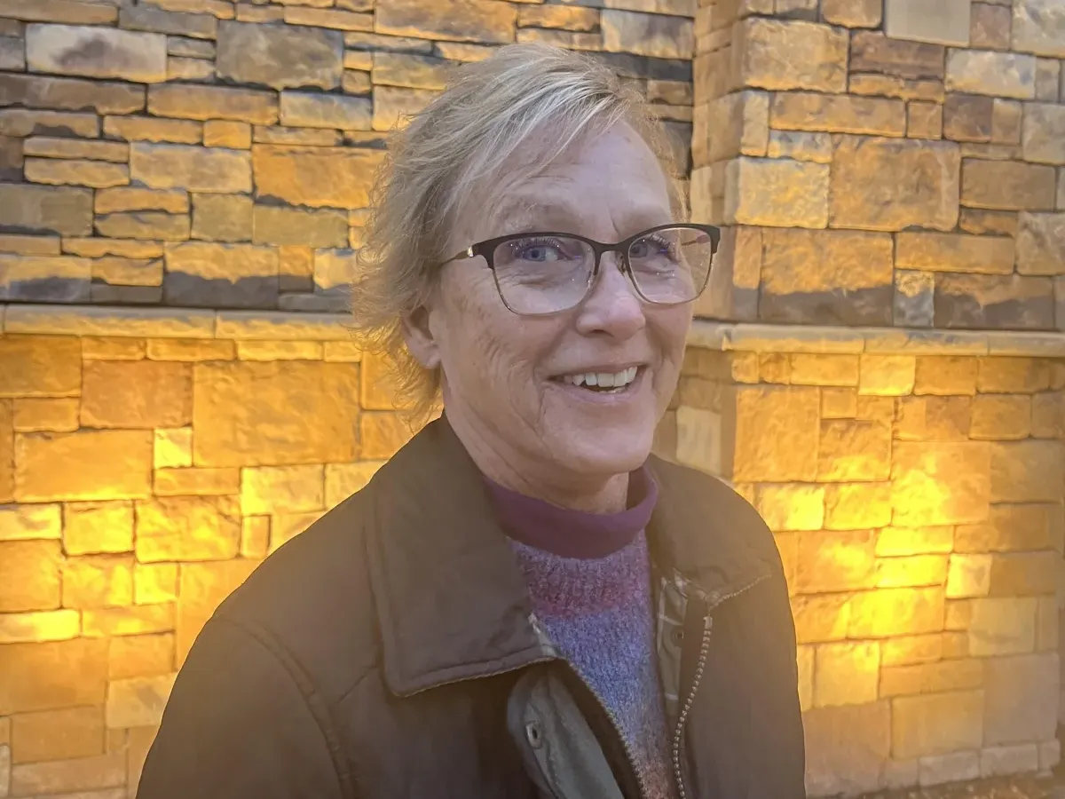 Smiling older woman with glasses and short hair stands by a stone wall lit with warm yellow lights.