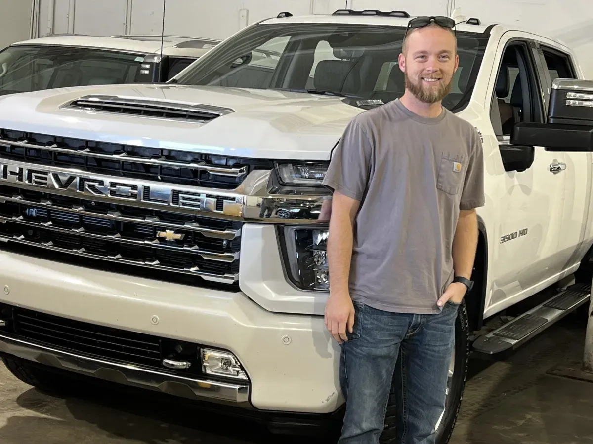 Man in gray shirt and jeans standing and smiling in front of a white Chevrolet Silverado pickup truck.