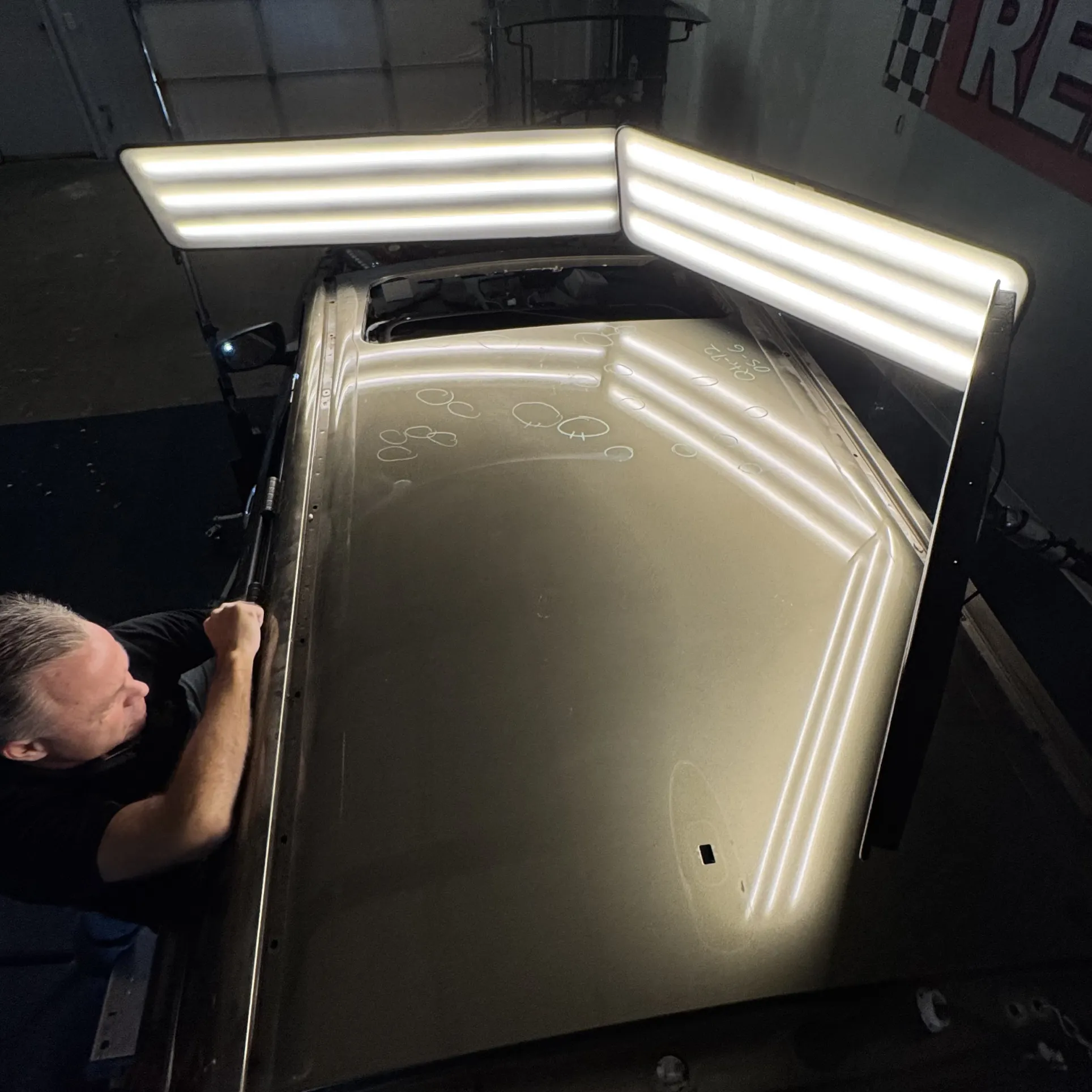 A person repairs a car roof with visible dents under bright inspection lights in a garage.
