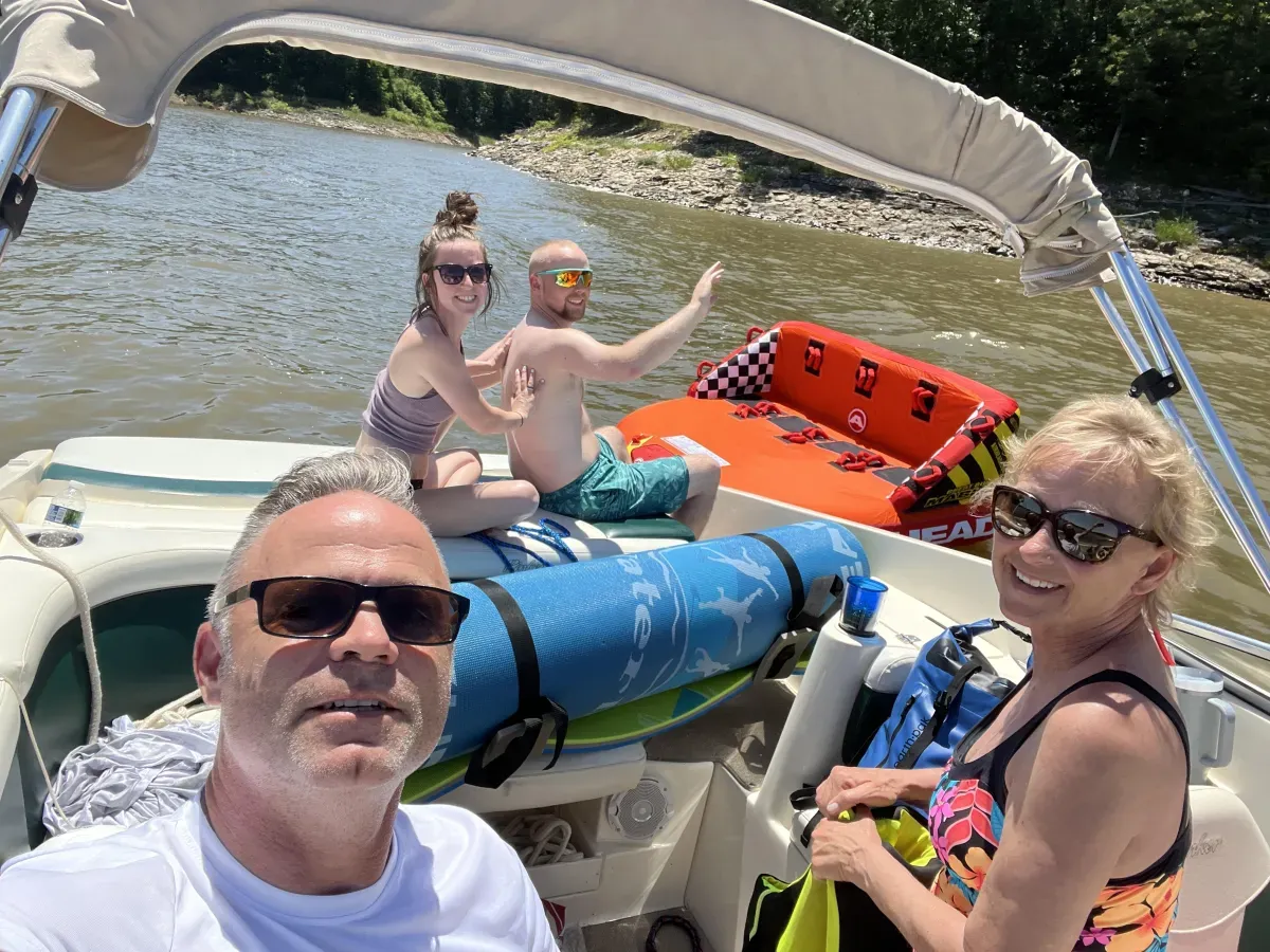 Four people smiling on a boat near the shore, with a lake and trees in the background on a sunny day.