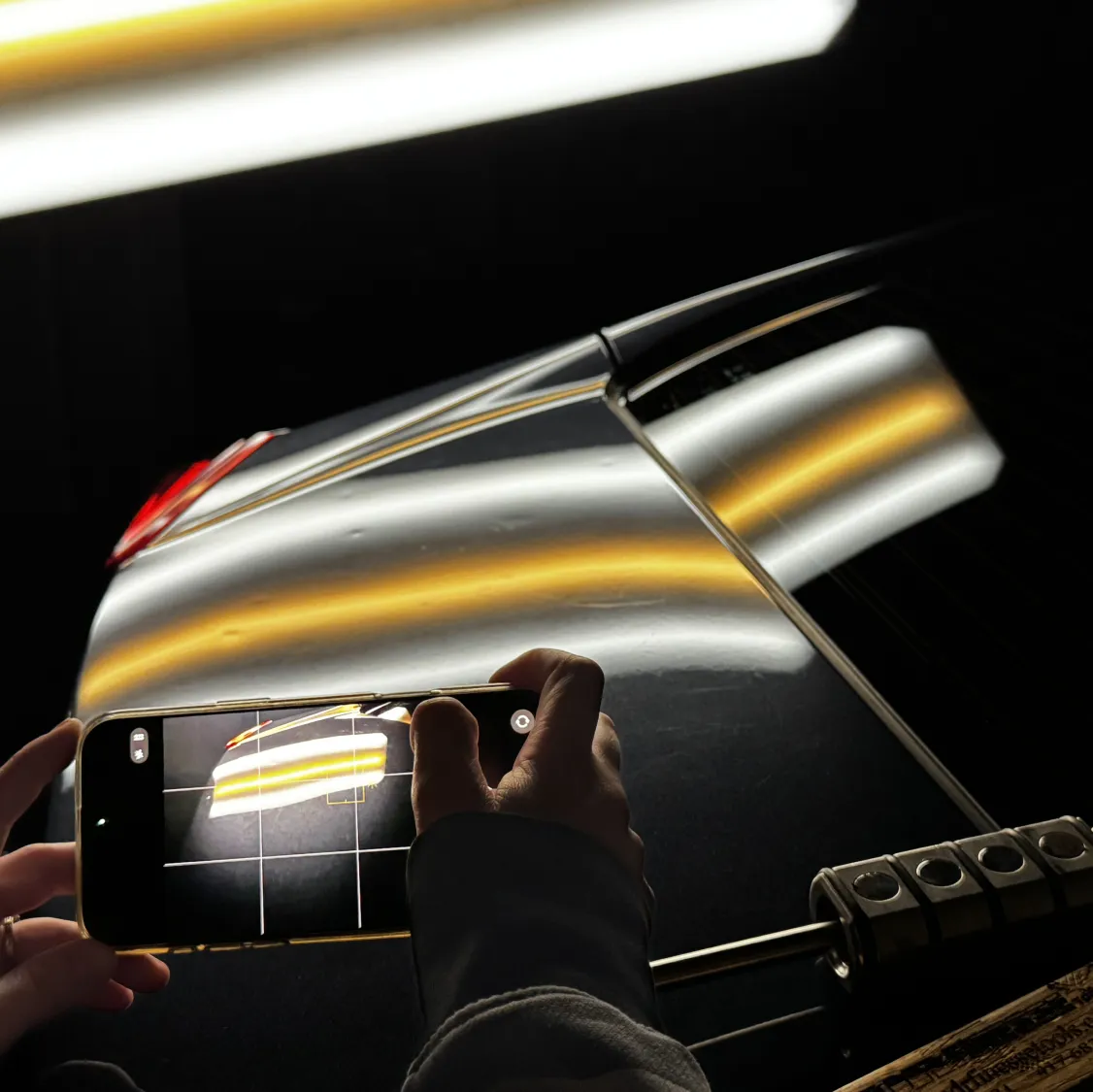 Hands holding a phone photographing yellow and white light reflections on a shiny black car surface.